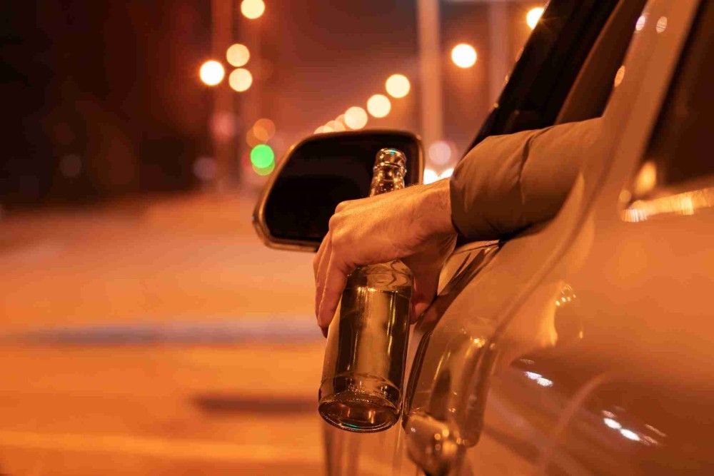 A hand holding a beer bottle out of a car window at night.